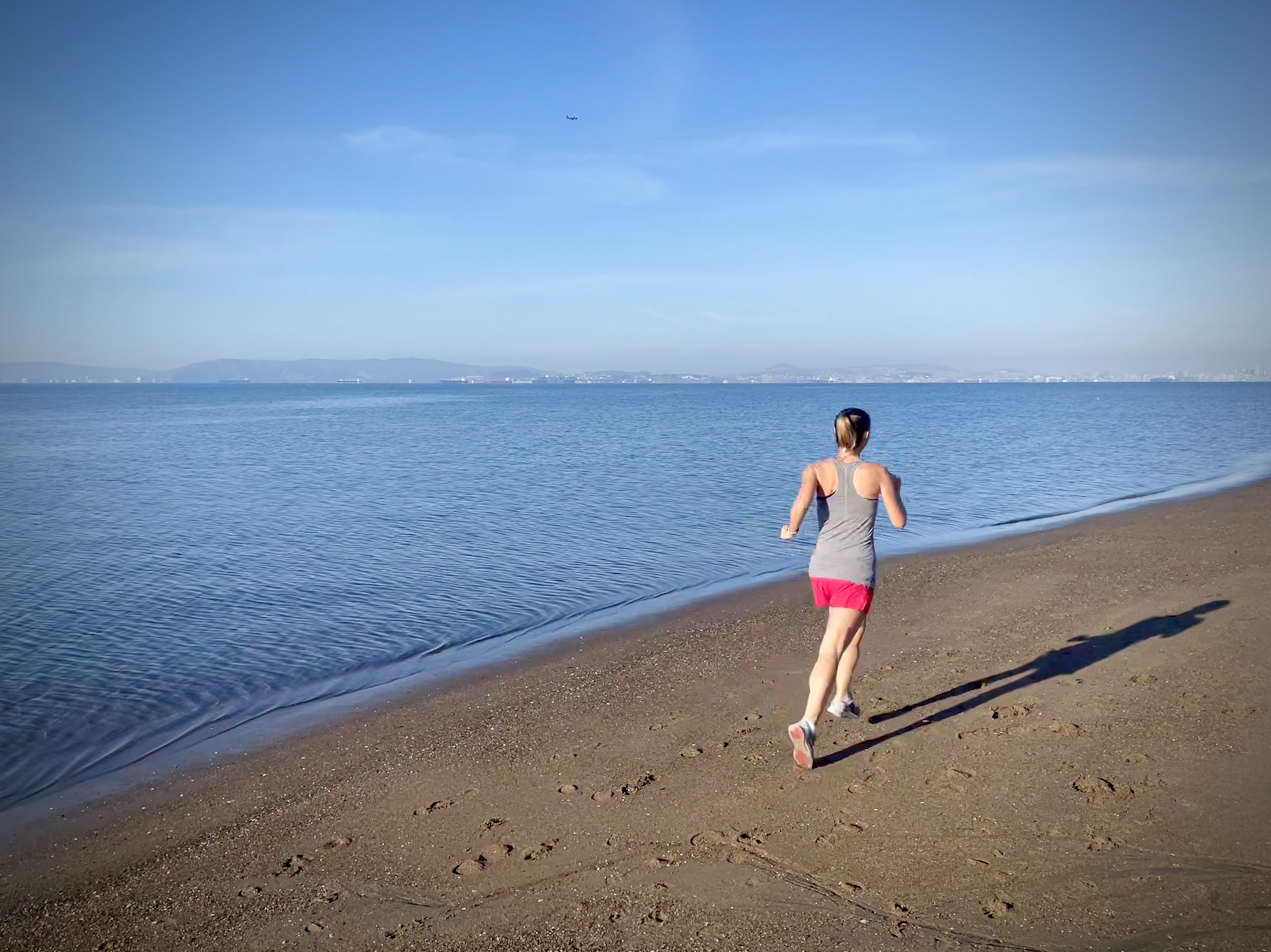Person running on beach towards horizon. It's morning, clear cky, blue ocean. You can see mountains far off in the distance.