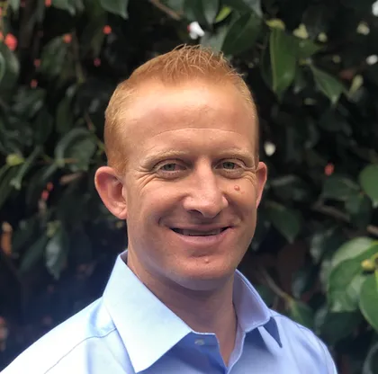 Professional portrait of Noah Merl from shoulders up. Fair skin, red hair, middle aged. Wearing light blue dress shirt in front of dark green foliage.